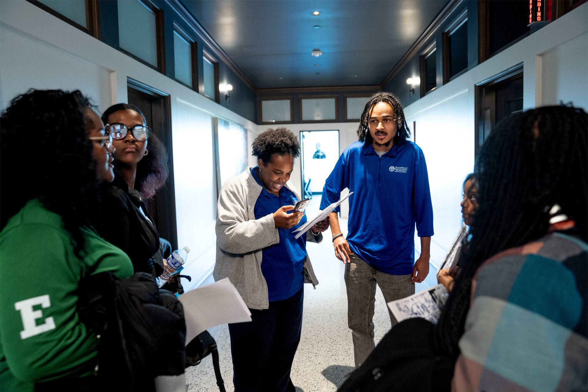Two GVSU staff members in blue Grand Valley State University polos review paperwork with a group of students gathered in a hallway.